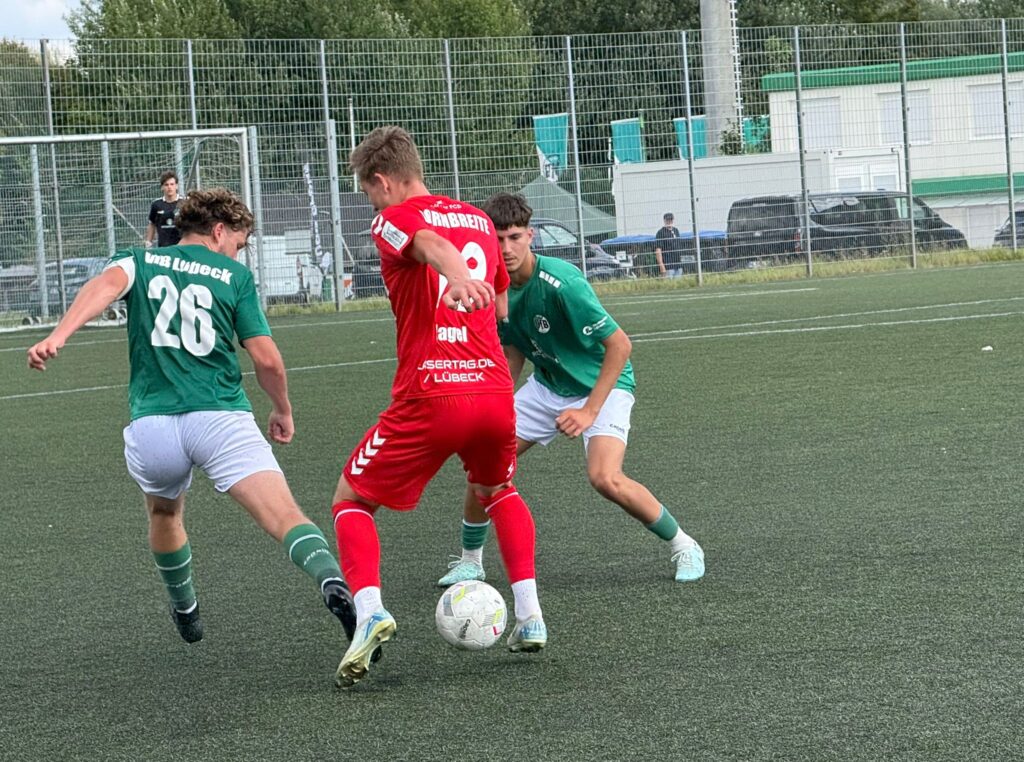 Drei Fußballer (Rot vs Grün) kämpfen um Ball auf Kunstrasen vor Tor und Zaun