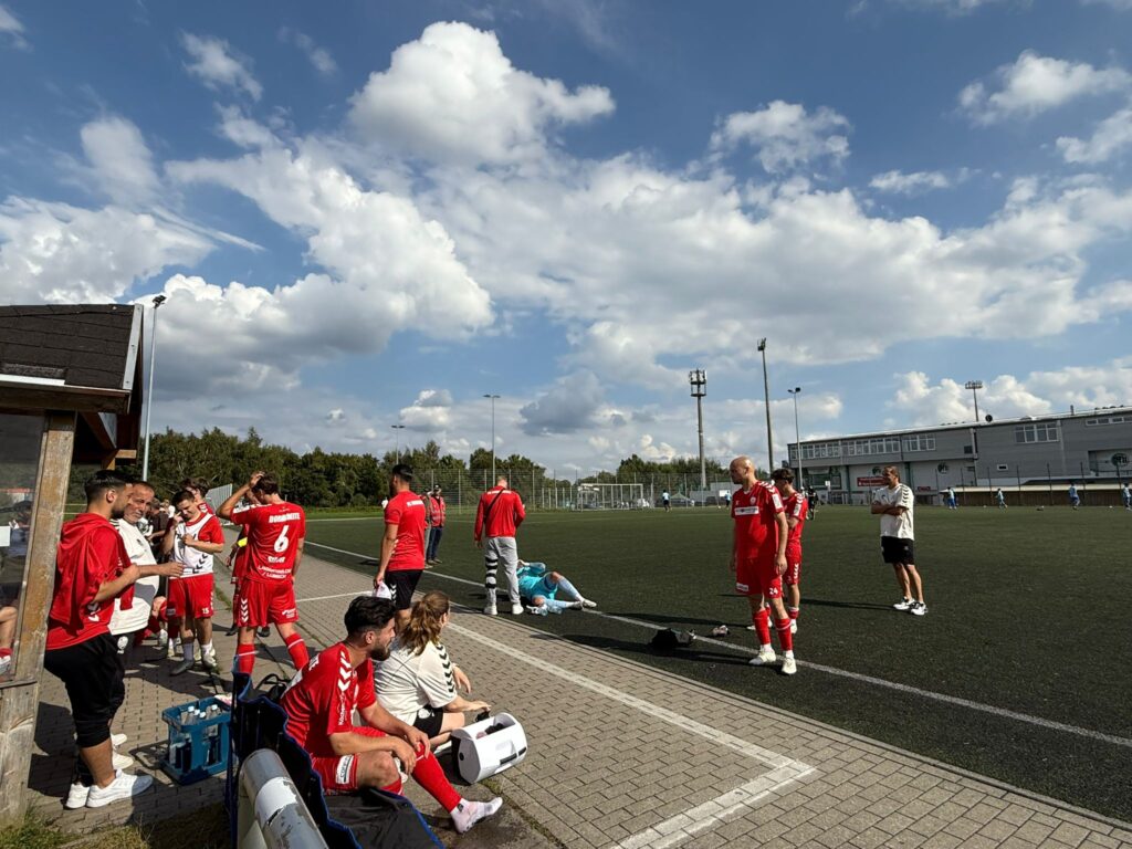 Fußballmannschaft in roten Trikots am Spielfeldrand bei sonnigem Himmel mit Wolken